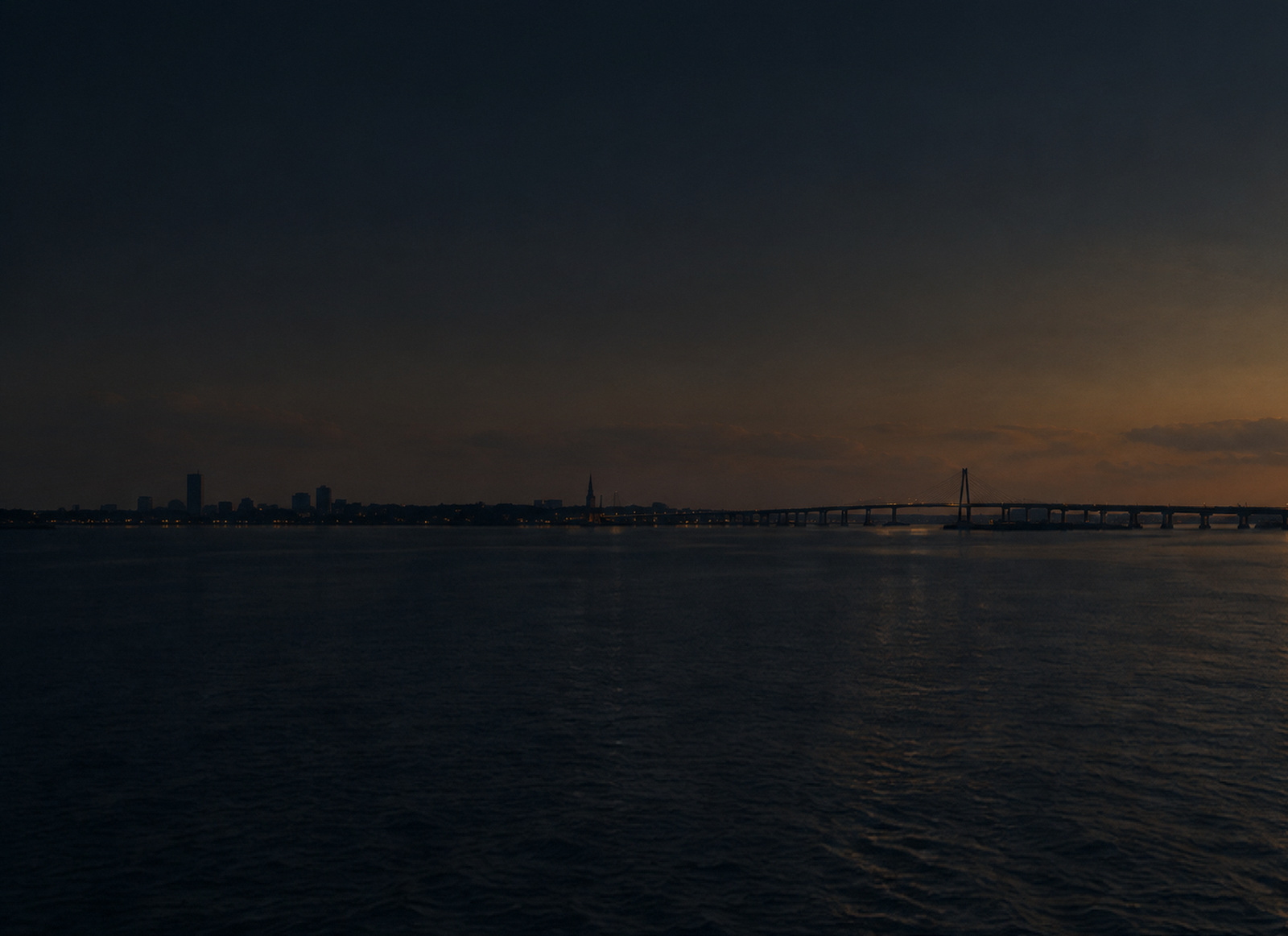Charleston harbor at dusk, the Ravenel Bridge and the peninsula skyline in silhouette against a low amber horizon.